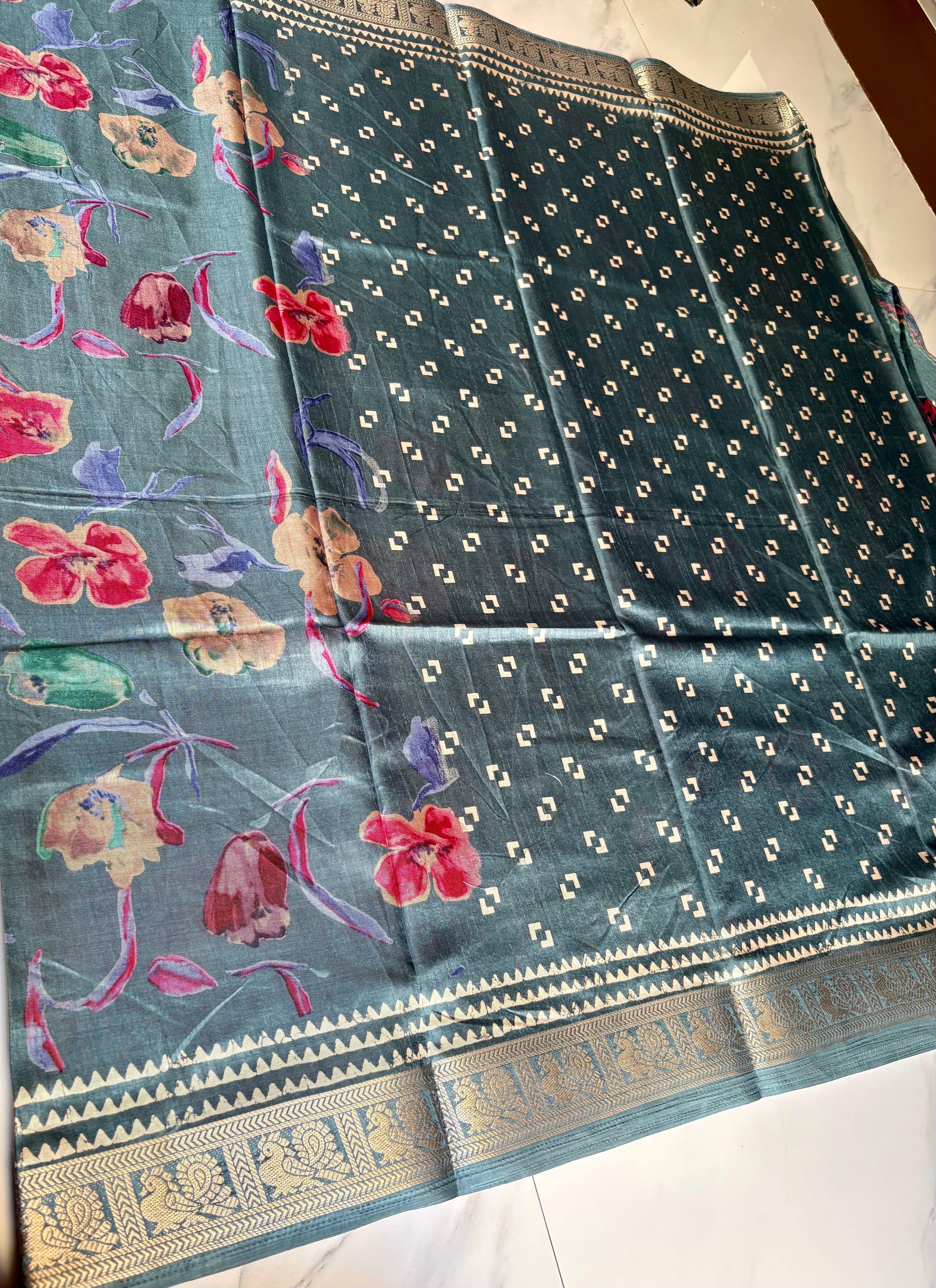 Elegant teal blue saree featuring vibrant multicolor floral prints that add a fresh and graceful charm. Finished with a rich golden woven zari border- Satin Tissue Saree